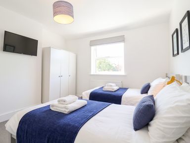 A bedroom with two single beds with blue throws and white towels a white wardrobe a wall-mounted tv and a window at Long View in Southwold