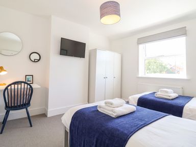 A bedroom with two single beds with blue throws and white towels a desk with a blue chair and two round mirrors a wardrobe and a wall mounted TV at Long View in Southwold