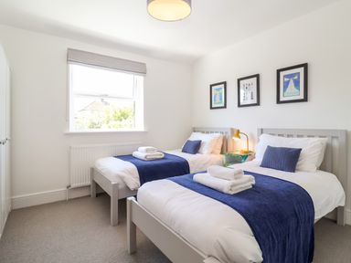 A bedroom with two single beds with blue throws and pillows a window and framed pictures on the wall at Long View in Southwold