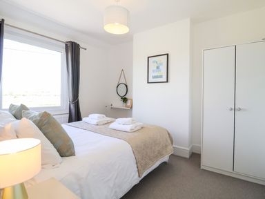 A bedroom with a bed linen and towels a wardrobe a window with curtains a wall mirror and a framed print at Long View in Southwold