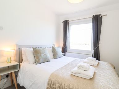 A bedroom with a bed with pillows and folded towels by a window with curtains at Long View in Southwold