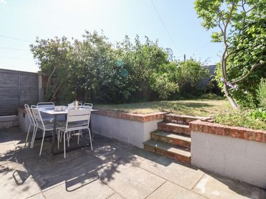 A backyard patio with a white metal table and chairs next to steps leading to a grassy garden with shrubs at Long View in Southwold