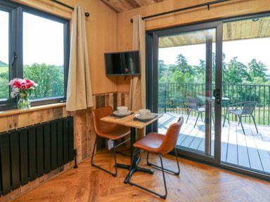 A dining area with a small table set for two and two brown chairs next to a window with flowers and a sliding door leading to a balcony with outdoor furniture at Hens Hideaway in Rhaedr Gwy near Rhayader