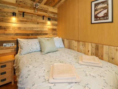 A bedroom with a bed with floral bedding two folded towels on the bed a bedside table wooden paneled walls and a framed picture at Hen's Hideaway in Rhaedr Gwy near Rhayader