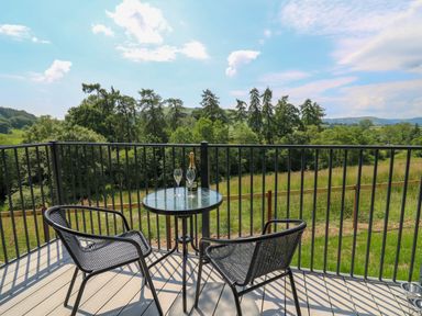 A balcony with a round glass table and two chairs overlooking green trees and fields at Hen's Hideaway in Rhaedr Gwy near Rhayader