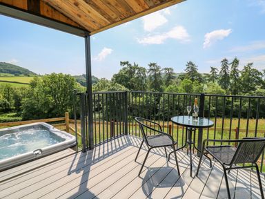 A balcony with a hot tub two chairs and a glass table with a bottle and glasses overlooking trees and hills at Hen's Hideaway in Rhaedr Gwy near Rhayader