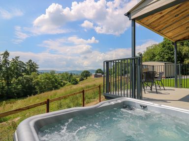An outdoor hot tub next to a covered patio with chairs and table overlooking a grassy hillside at Hen's Hideaway in Rhaedr Gwy near Rhayader
