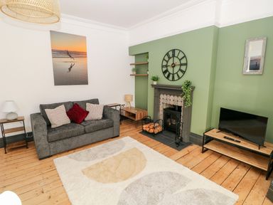 A living room with a grey sofa and cushions a fireplace with a clock above a TV on a wooden stand and a rug on a wooden floor at 36A Mary Street in Porthcawl