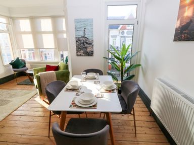 A dining area with a white table set with plates and glasses surrounded by gray chairs next to a green armchair and a floor lamp at 36A Mary Street in Porthcawl
