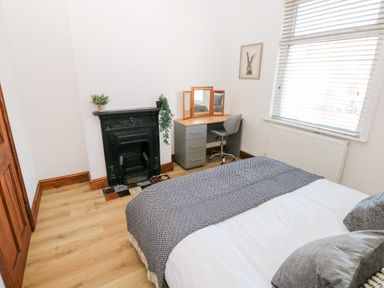 A bedroom with a bed, a desk with a mirror and chair, a black fireplace and a window with blinds at 36A Mary Street in Porthcawl