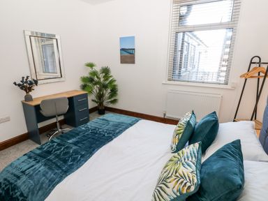 A bedroom with a bed with pillows and a blue throw blanket a desk with a chair and a mirror a plant and a window with blinds at 36A Mary Street in Porthcawl