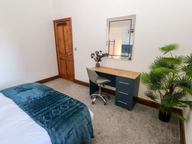 A bedroom with a bed, a desk with drawers and a chair, a mirror on the wall, a wooden door, and a potted plant at 36A Mary Street in Porthcawl