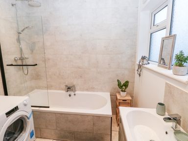 A bathroom with a bathtub and shower, a washing machine, a sink with a faucet, a window with plants and a mirror on the sill at 36A Mary Street in Porthcawl