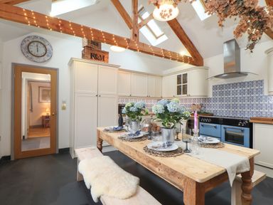 A kitchen with a wooden dining table set with plates flowers and glassware under wooden beams with string lights and tiled backsplash at The Riblows Cape Cornwall in Bollowal near St Just