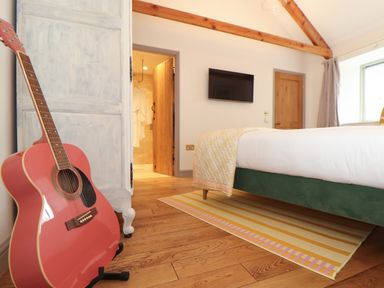 A bedroom with a bed a wall-mounted tv a wooden door a striped rug a guitar on a stand and an open door revealing a bathroom at The Riblows Cape Cornwall Bollowal near St Just