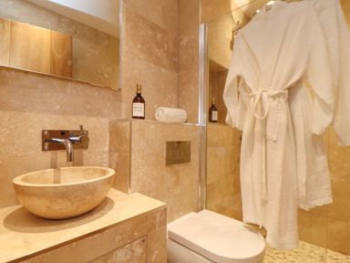 A bathroom with a stone sink, toilet, white robe hanging, hand wash bottle, and rolled towel at The Riblows, Cape Cornwall in Bollowal near St Just