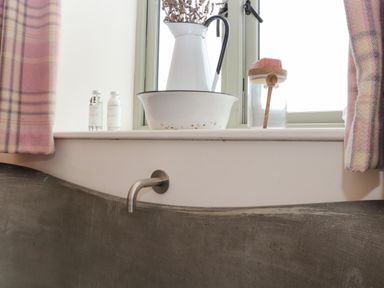 A bathroom window sill with bottles a white pitcher with dried flowers and a soap jar above a concrete bathtub at The Riblows Cape Cornwall Bollowal near St Just