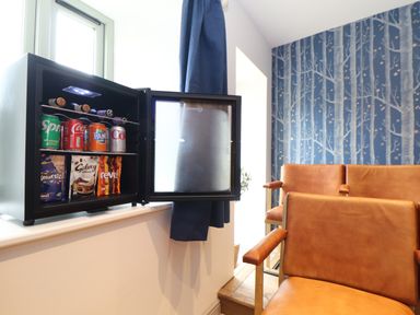 A small refrigerator with drinks and snacks inside next to brown leather chairs and a wallpapered wall with tree design at The Riblows Cape Cornwall Bollowal near St Just
