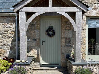 A front door with a wooden porch and wreath surrounded by stone walls and plants at The Riblows Cape Cornwall Bollowal near St Just