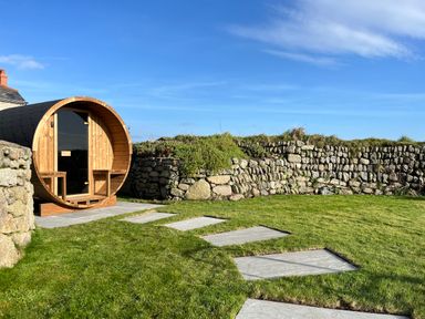 A circular wooden structure with a glass door on a stone patio in a grassy yard with a stone wall at The Riblows Cape Cornwall