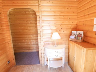 An interior corner of a wooden cabin with an arched doorway a white table lamp on a small white drawer unit and a brochure box on a wooden cabinet at 64 Acorn Lodge Kenwick Park in Louth