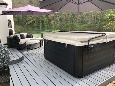 An outdoor deck with a hot tub a wicker sofa set with cushions and two patio umbrellas surrounded by trees at Glamorgan House in Cardiff