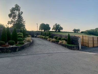 A driveway with stone walls and landscaped plants leading to a gate with trees and grass fields in the background at Glamorgan House in Cardiff