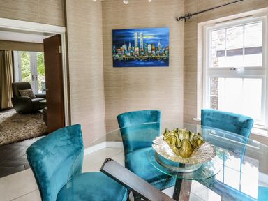 A dining area with a glass round table and four blue chairs a decorative bowl on the table and a cityscape painting on the wall at Glamorgan House in Cardiff