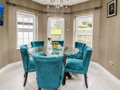 A dining room with a round glass table and six teal chairs near three large windows at Glamorgan House in Cardiff