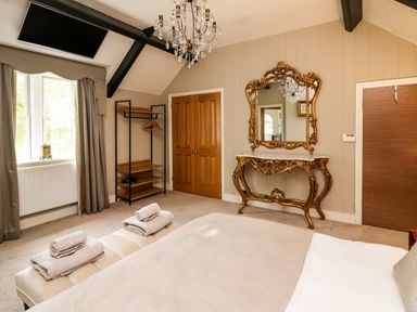 A bedroom with a bed, chandelier, ornate mirror and console table, wooden doors, window with curtains, and a shelving unit at Glamorgan House in Cardiff