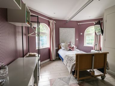 A bedroom with a single bed with pillows and a blanket a wall-mounted television two arched windows curtains a metal frame with hangers and shelves and a patterned rug at Glamorgan House in Cardiff