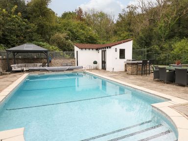 An outdoor swimming pool with steps and seating areas including a gazebo and chairs near a small building surrounded by trees at Glamorgan House in Cardiff