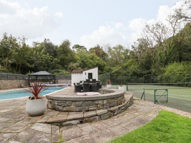 An outdoor area with a swimming pool, patio dining set, planters, and a tennis court surrounded by trees