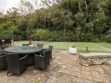 An outdoor patio area with a round table and wicker chairs next to a fenced tennis court surrounded by trees at Glamorgan House in Cardiff
