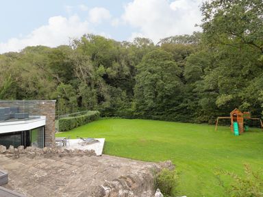 A backyard with a patio area and lounge chairs next to a grassy field with a children's playset and trees in the background at Glamorgan House in Cardiff