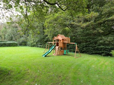 A wooden playground set with swings slide and climbing wall on a grassy lawn surrounded by trees at Glamorgan House in Cardiff