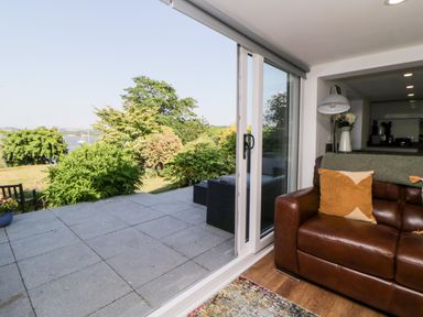 A living room with a brown leather sofa next to large sliding glass doors opening to a patio and garden at Riverview @ 1 Brunel View in Saltash