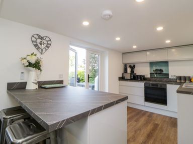 A kitchen with a black countertop island and bar stools a wall clock and flowers on the counter built in oven and white cabinets at Riverview @ 1 Brunel View Saltash
