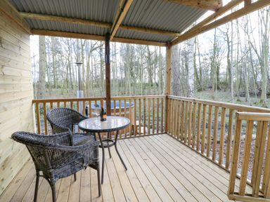 A wooden covered porch with two wicker chairs a glass top table with a bottle and glasses and a hot tub outside at The Leveret in Eppleby