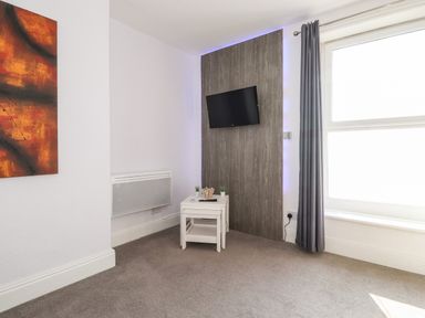 A corner of a room with a wall mounted television above two small white tables near a window with grey curtains at Apartment 9 Beaconsfield House in Bridlington