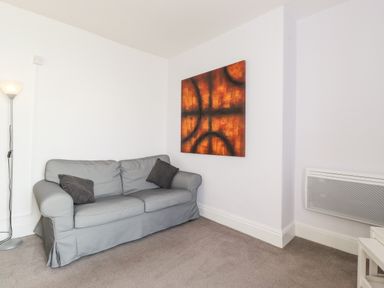 A living room with a gray sofa two dark cushions an orange abstract painting a floor lamp and a wall heater at Apartment 9 Beaconsfield House in Bridlington
