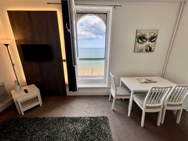 A room with a window overlooking the beach and sea a wall-mounted TV a white dining table with three chairs and a floor lamp at Apartment 9 Beaconsfield House Bridlington