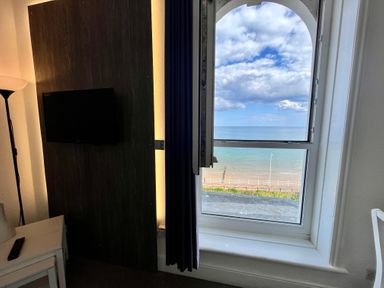 A room with a wall-mounted TV a floor lamp a white table and an open window showing a view of the beach and sea at Apartment 9 Beaconsfield House in Bridlington