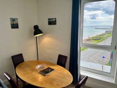 A dining room with a wooden table and four chairs near a window overlooking a coastal road at 7 Beach View @ Beaconsfield House in Bridlington