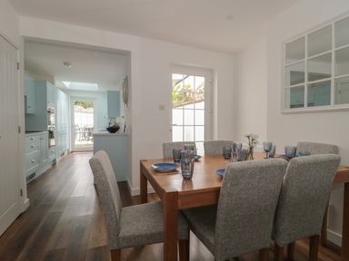 A dining area with a wooden table and six grey upholstered chairs next to a kitchen with light blue cabinets at Quay Cottage - Sea View in Poole