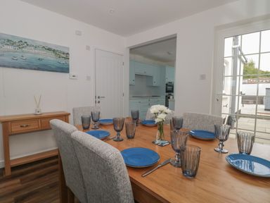 A dining room with a wooden table set with blue plates and glasses gray upholstered chairs a wooden side table with a diffuser painting on the wall and a view into a kitchen with blue cabinets at Quay Cottage Sea View in Poole
