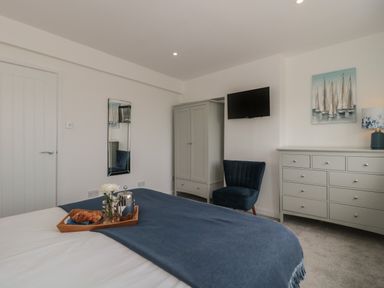 A bedroom with a bed covered by a blanket with a tray holding croissants and a teapot a blue chair a wardrobe a dresser a mounted TV and a painting of sailboats at Quay Cottage - Sea View in Poole