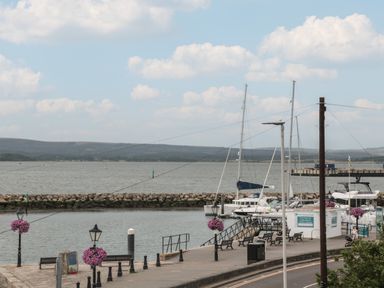 A dock with benches, lampposts with hanging flower baskets, boats docked in the water, and hills in the background at Quay Cottage Sea View in Poole