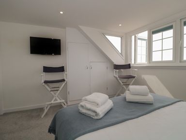 A bedroom with two folded towel sets on a bed with a blue blanket two chairs near windows and a wall-mounted TV at Quay Cottage - Sea View in Poole