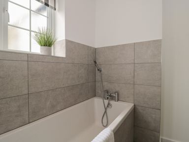 A bathroom with a bathtub gray tiled walls a window with a potted plant and a handheld shower at Quay Cottage - Sea View in Poole